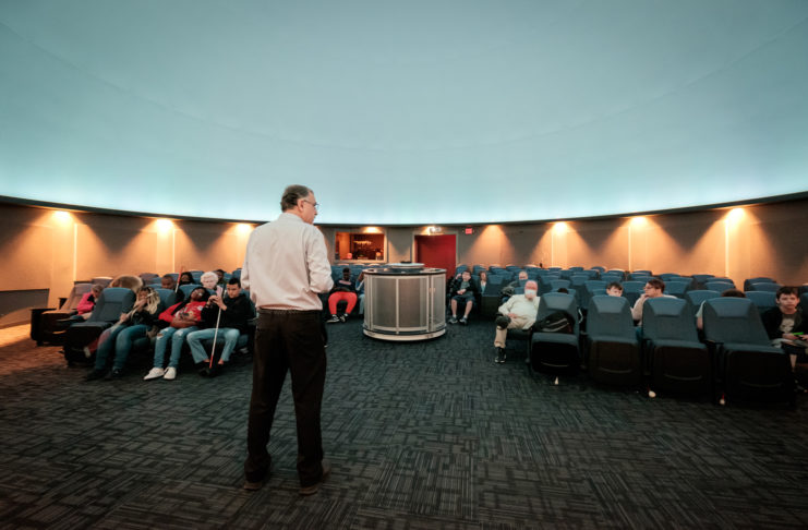 A man stands in front of a group of students seated in a planetarium