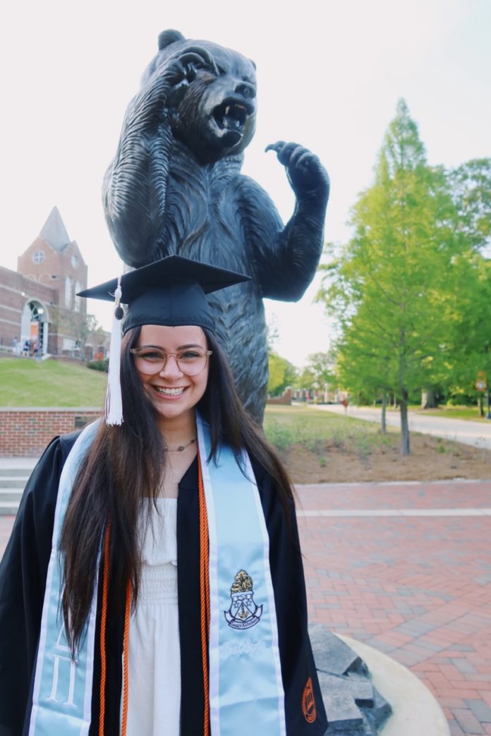 A young woman in a graduation cap and gown stands in front of a bear statue