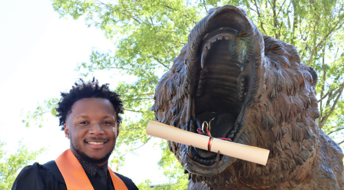 Graduate Spotlight: Willie Fulks A young man wearing a graduation gown and orange stoll stands next to a bear statue. The statue has a rolled up piece of paper, like a diploma, in its mouth.