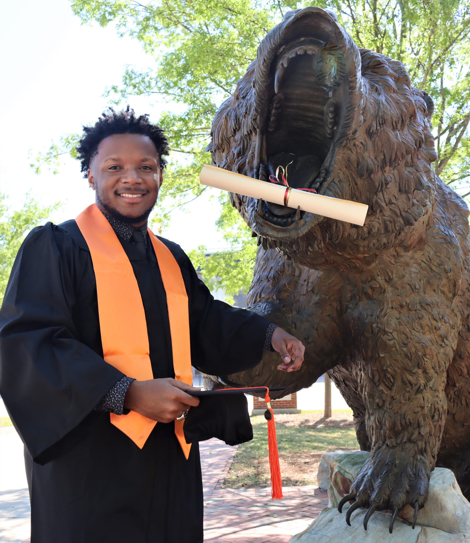 A young man wearing a graduation gown and orange stoll stands next to a bear statue. The statue has a rolled up piece of paper, like a diploma, in its mouth.