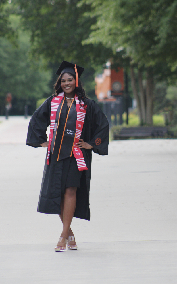 A young woman wearing a graduation cap, gown and cords stands with her hands on her hips