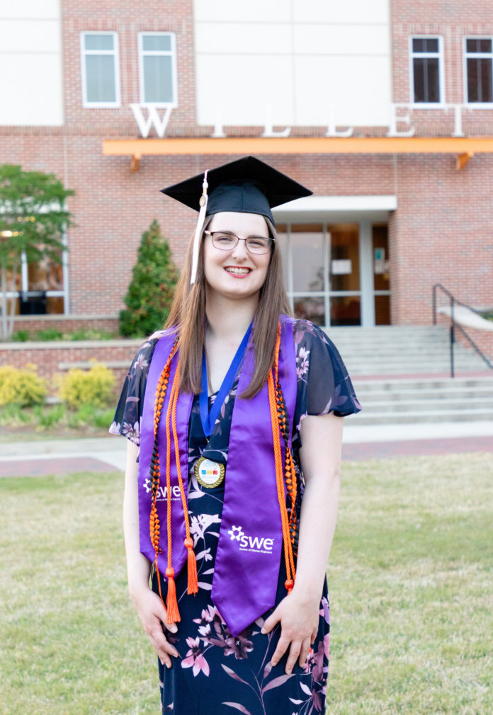 A young woman wears a graduation cap and honors cords while standing in front of a building