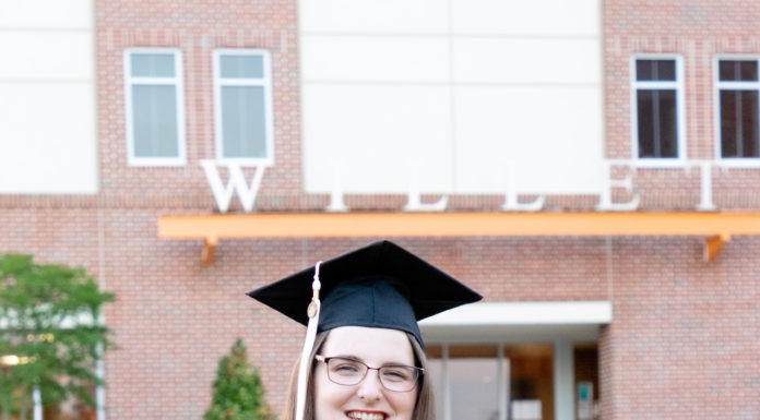 Graduate Spotlight: Emily Wilbourn A young woman wears a graduation cap and honors cords while standing in front of a building