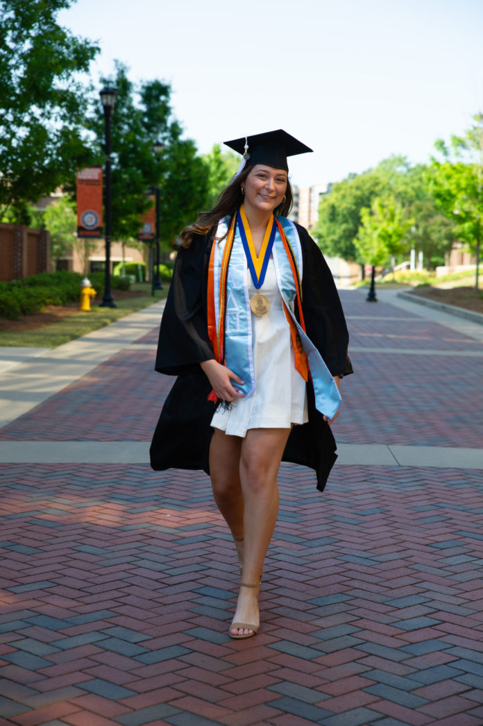 A young woman wearing a graduation cap, gown and cords walks down a brick pathway