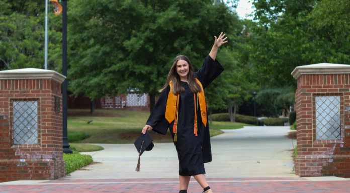 Graduate Spotlight: Mallory Green A young woman wearing a graduation gown spreads her arms wide. There is a graduation cap in her left hand.