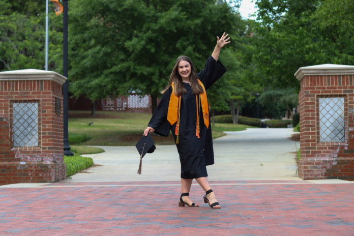A young woman wearing a graduation gown spreads her arms wide. There is a graduation cap in her left hand.