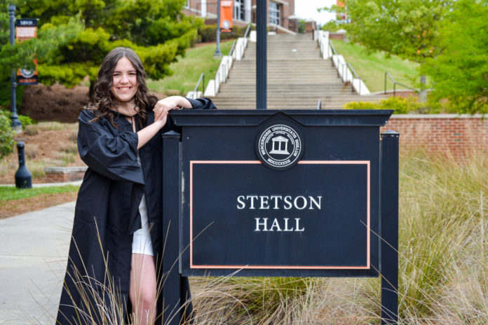 A young woman in a graduation gown stands next to a sign a sign for Stetson Hall
