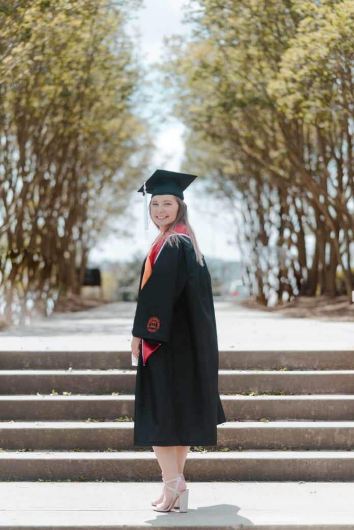 A young woman wearing a graduation cap and gown stands outside in front of steps
