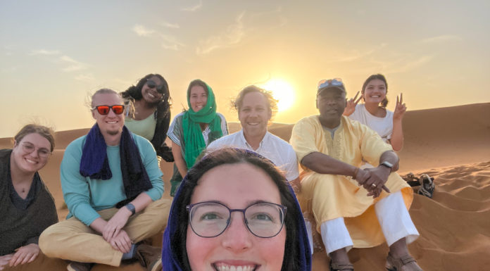 Mercerians find common ground between Christians and Muslims while studying in Morocco Group of people sitting on a sand dune with the sunset in the background, smiling at the camera. Everyone is casually dressed, some wearing scarves, in a desert setting.