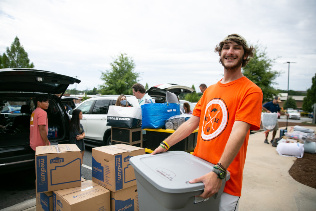 A young man wearing an orange shirt carries a large gray container. Cars and boxes are in the background