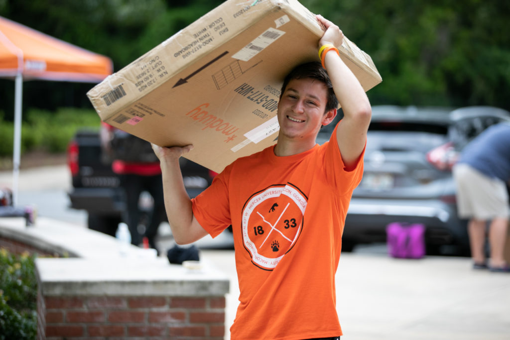 A young man wearing an orange shirt carries a box over his head