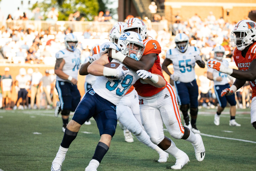 A football player in a white and blue uniform is tackled by a player in an orange uniform.