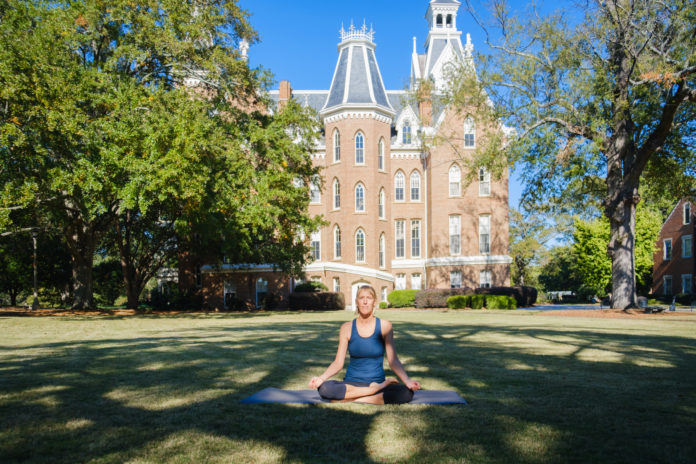 A woman sits cross-legged on a yoga mat in front of Mercer's administration buliding