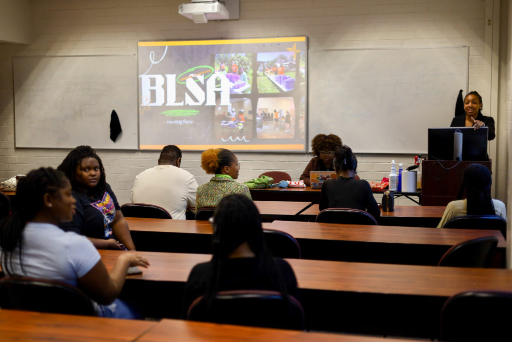 A young woman stands at a podium in front of students sitting at desks. the screen behind here reads BLSA
