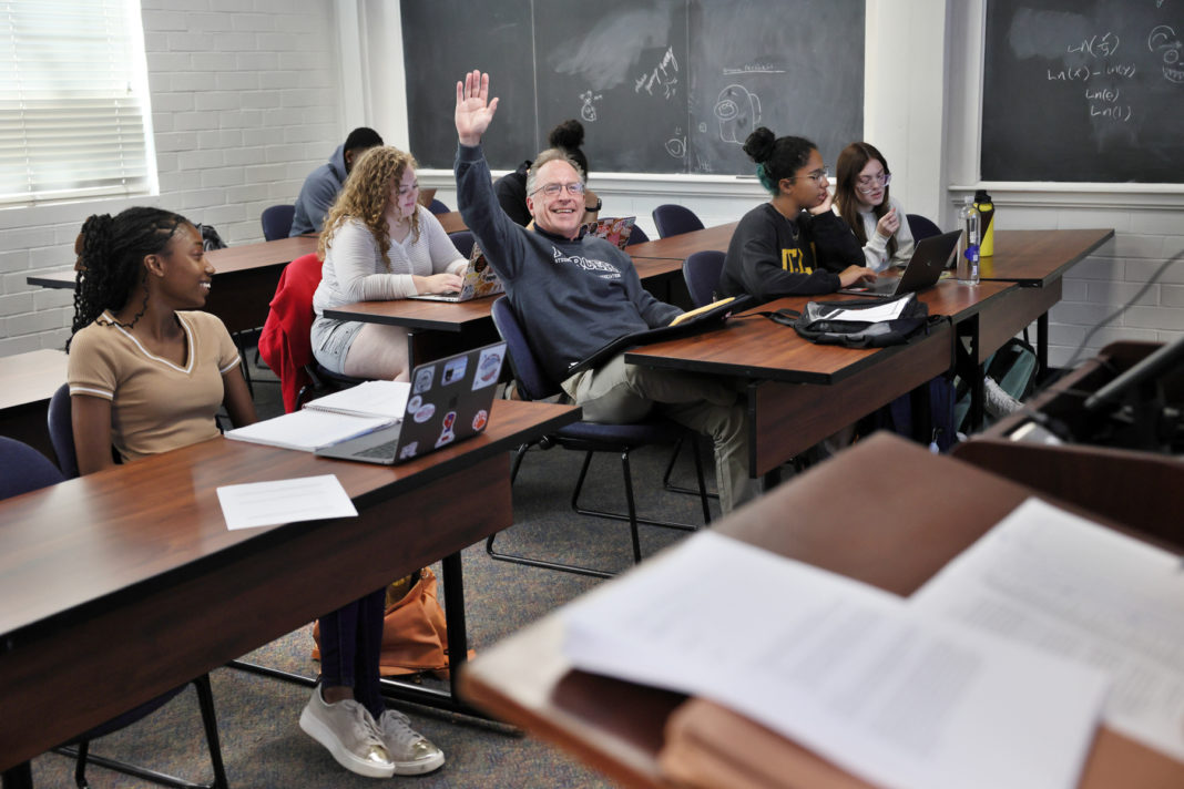 A man wearing a gray Mercer sweatshirt sits at a desk surrounded by other students and raises his hand