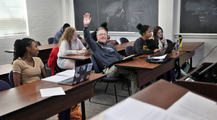 Mercer dean of students and SGA president trade places for a day A man wearing a gray Mercer sweatshirt sits at a desk surrounded by other students and raises his hand