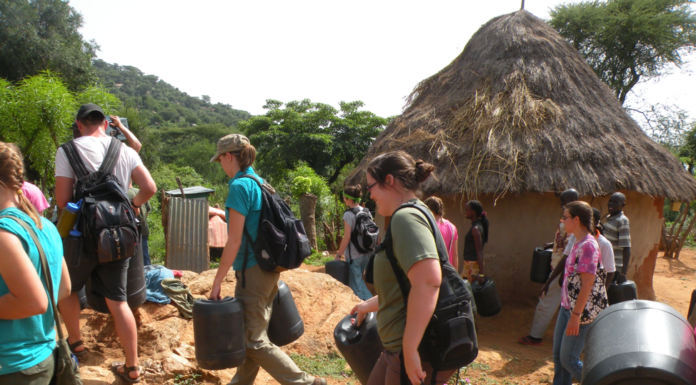 Lessons in thankfulness learned from Mercer On Mission people in western clothing carry large jugs past a mud house with a thatch roof