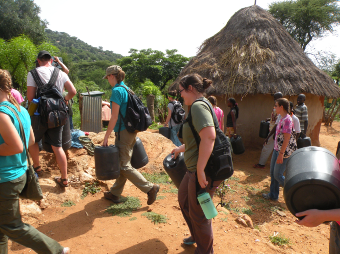 people in western clothing carry large jugs past a mud house with a thatch roof