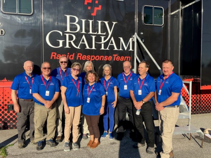 Ten people wearing blue shirts and khaki pants stand in front of a large vehicle that says 