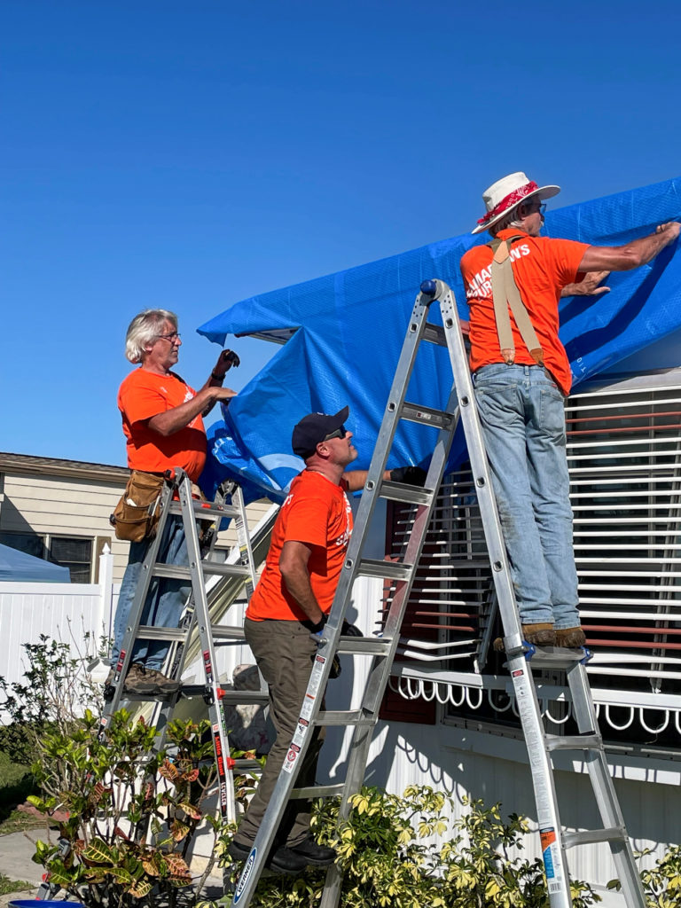 Three men on ladders put a blue tarp on a house