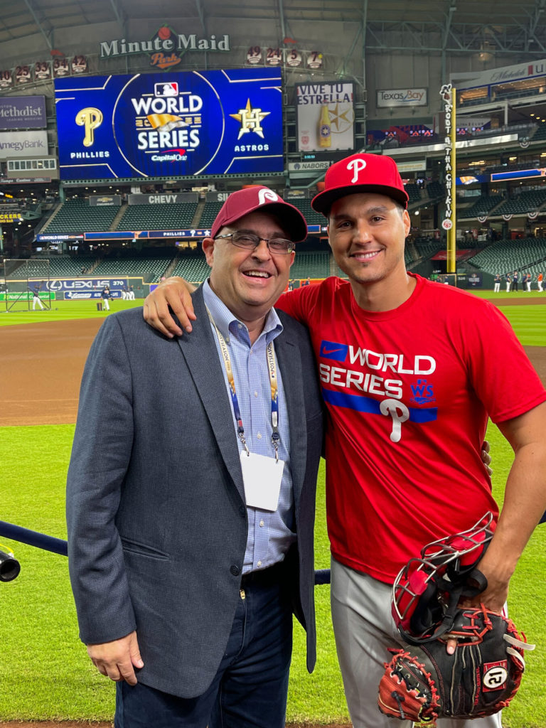 Dos hombres se paran uno al lado del otro frente a un campo de béisbol.  El logotipo de la Serie Mundial se muestra en la pantalla grande detrás de ellos.