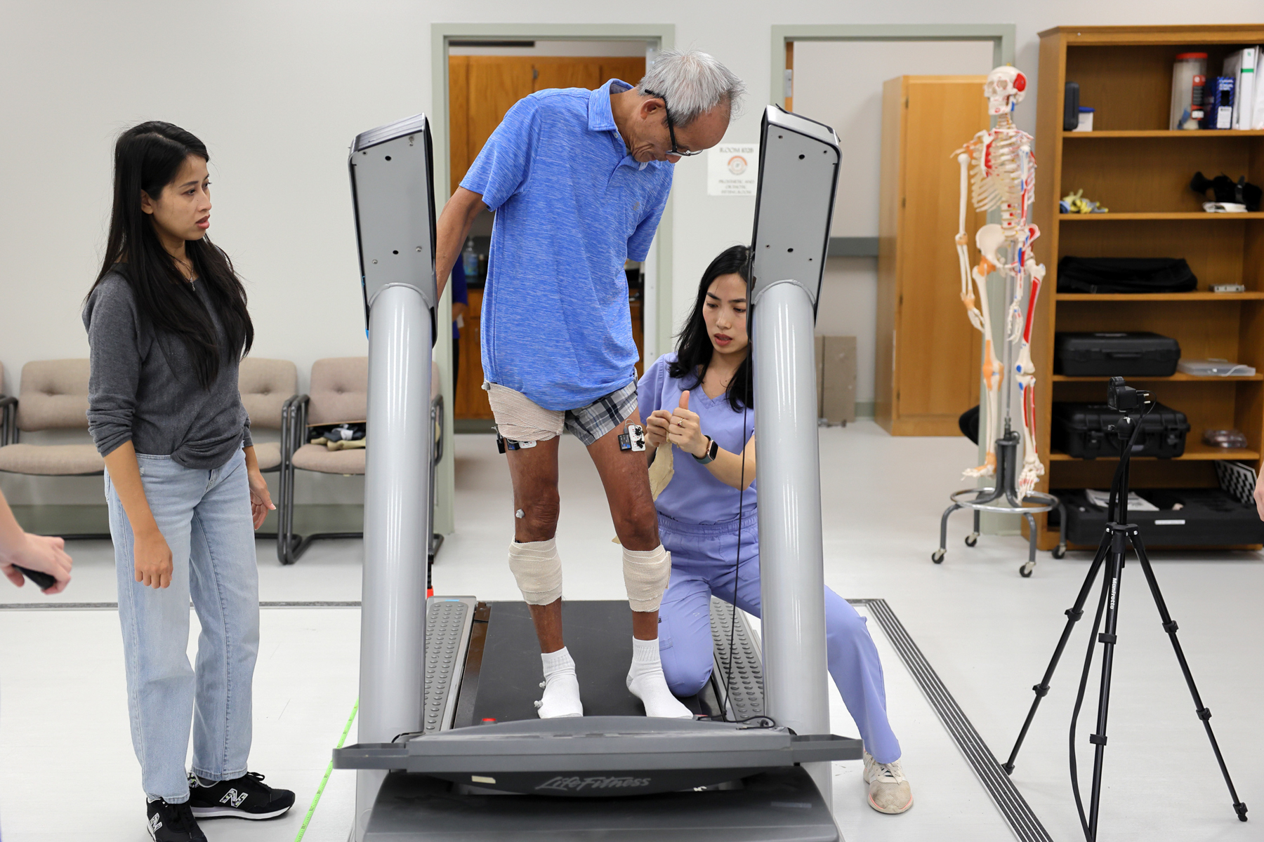 A man stands on a treadmill while a woman in blue scrubs places a bandage on his leg