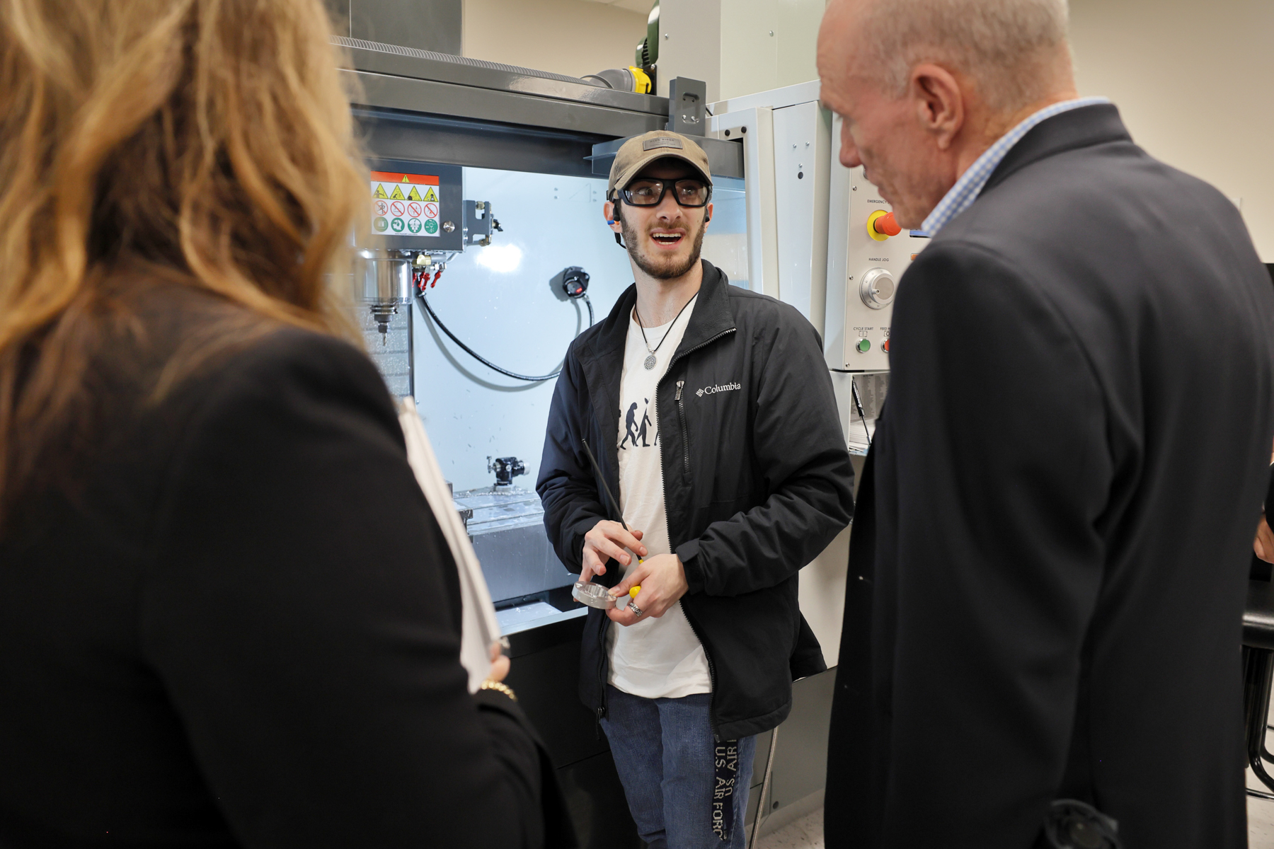 A man wearing safety glasses speaks to a man and a woman in front of a machine