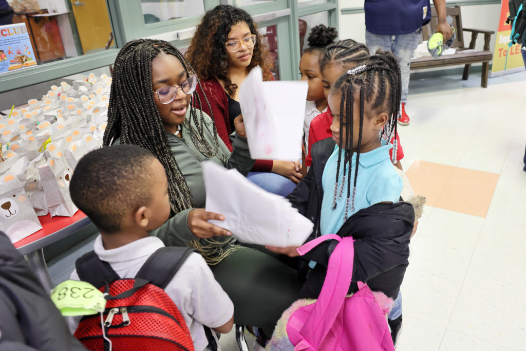 Two seated college students are surrounded by four elementary school students who are showing the college students their papers.