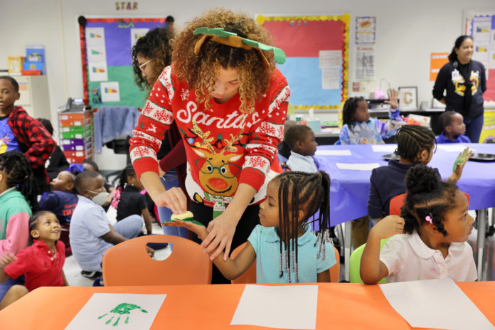 A college student wearing a red sweater with a reindeer on it, using a sponge to put green paint on the palm of an elementary school student's hand.