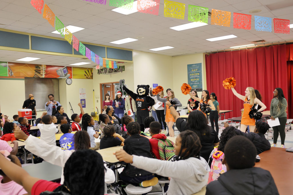 A room full of children sitting at tables. At the front of the room, a bear mascot and cheerleaders lead the students in a cheer.