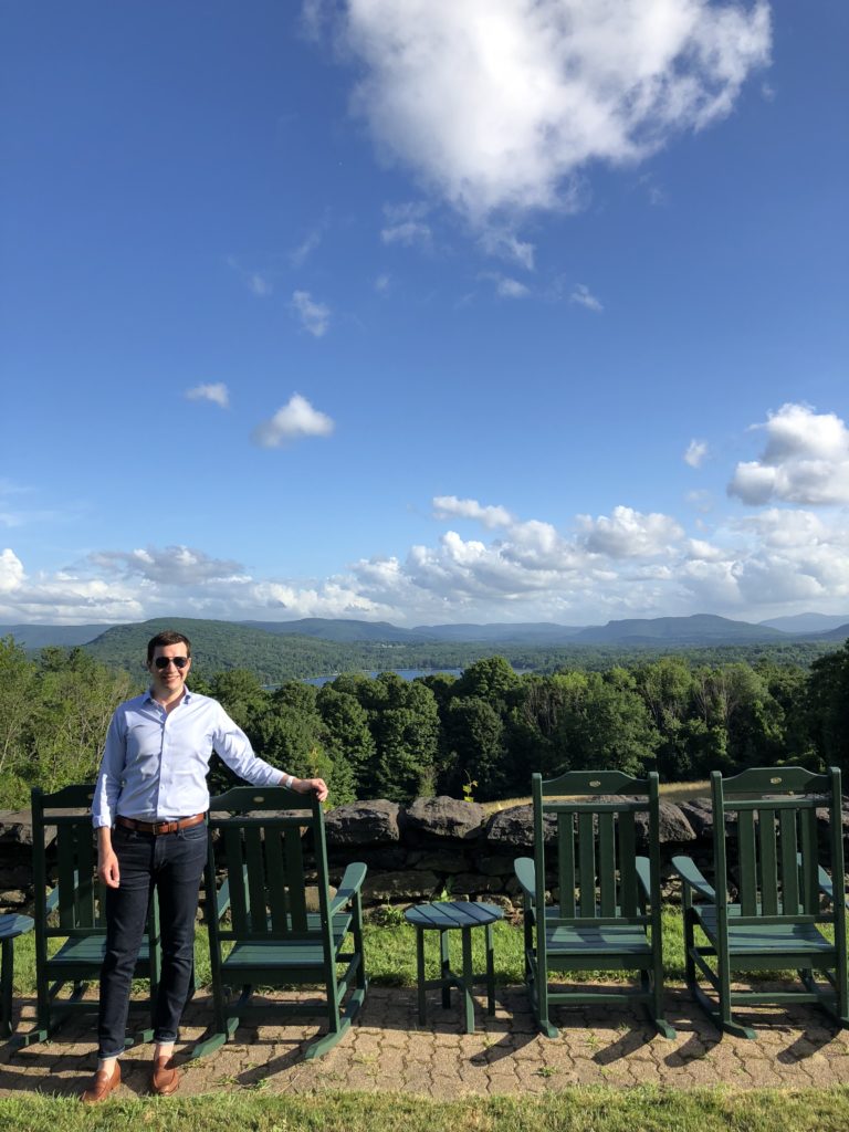 Alumnus Andrew Lane is shown in New England with green rocking chairs and mountains behind him.