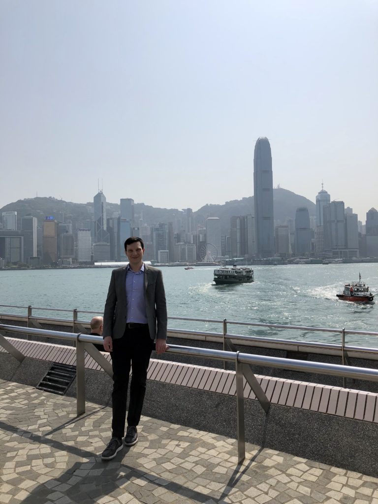 Alumnus Andrew Lane is shown in Hong King, with boats on the water and the city skyline behind him.