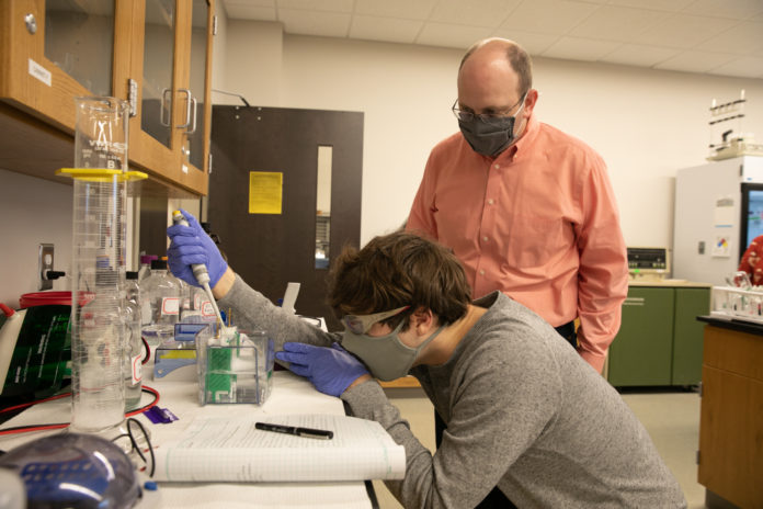 A male professor looks on while a male student pipes a liquid into a container