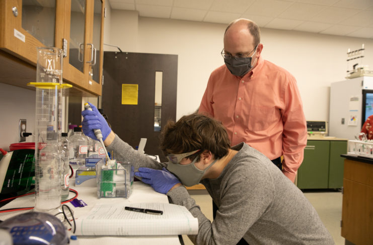 A male professor looks on while a male student pipes a liquid into a container