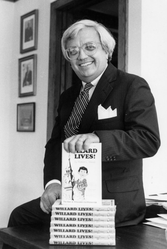 Bob Steed is wearing a suit and sitting on a table with his hand atop a stack of books. the cover, says Willard Lives! and pictures a cartoon young man next to the administration tower