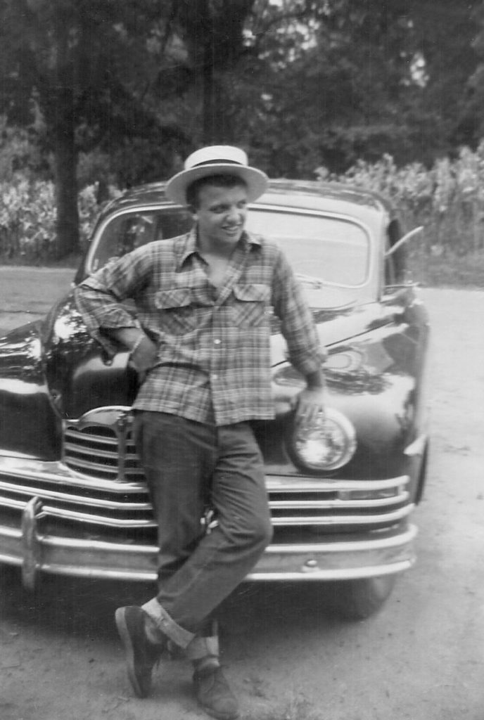 a young man wearing an untucked checkered shirt and jeans stands in front of an old car. the photo is black and white
