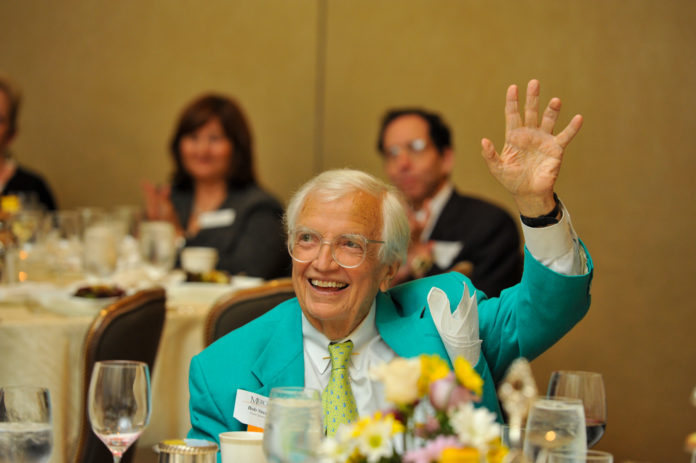bob steed sits at a table decorated for a luncheon. he looks up and is waving his hand smiling.