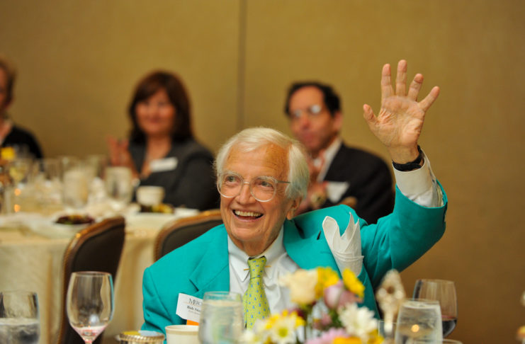 bob steed sits at a table decorated for a luncheon. he looks up and is waving his hand smiling.