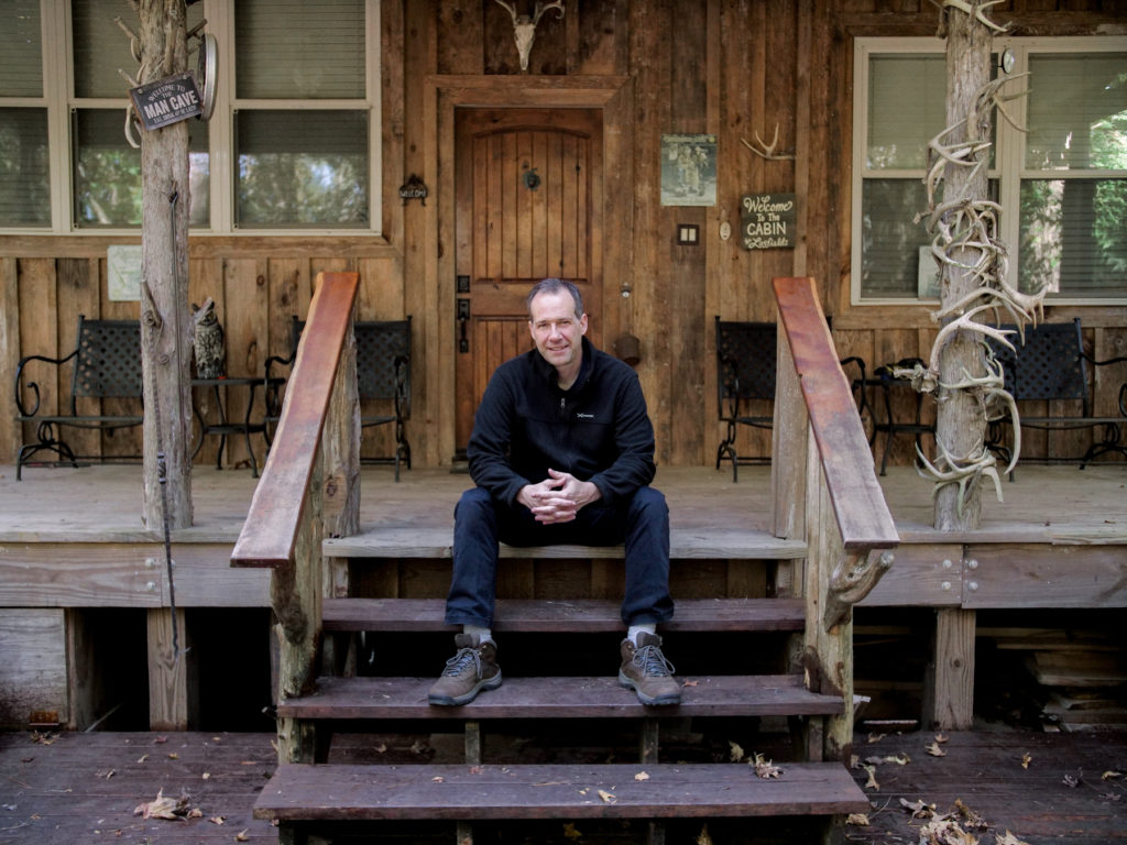 Dr. Christopher Schmitz, wearing a black pullover and pants, along with hiking boots, sits on the front steps of a cabin.