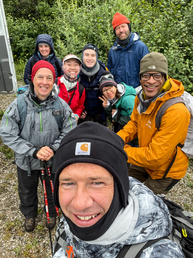 A group of eight people wearing hiking and rain gear take a selfie in the woods.