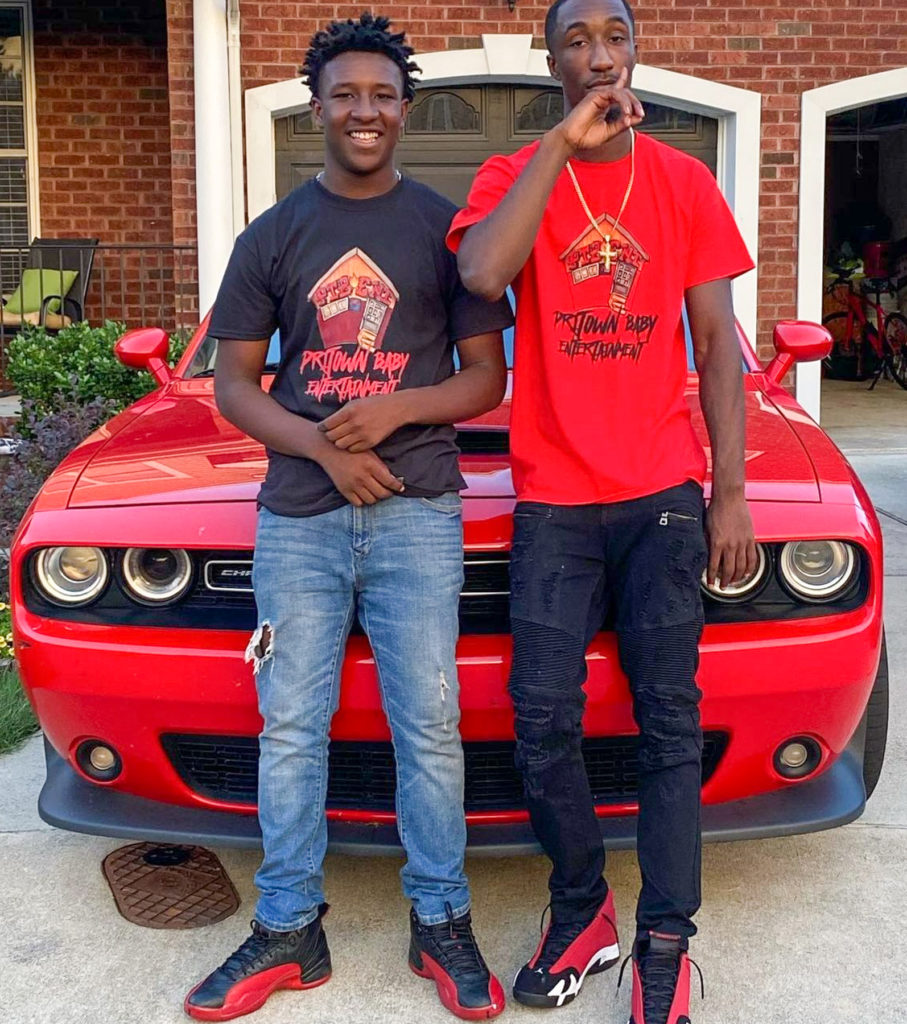Two young men wearing shirts that say Pritown Baby Entertainment stand in front of a red sports car.