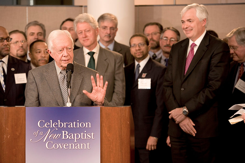 Jimmy Carter speaks at a podium that has a sign that says "Celebration of a New Baptist Covenant." Bill Clinton is behind Carter and William D. Underwood is to his right.