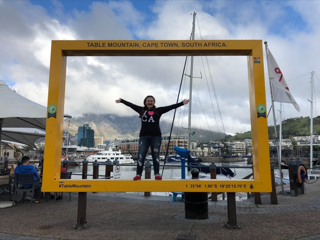 Johna Wright is shown in front of a Cape Town photo op with mountains and ships in the backgroun.