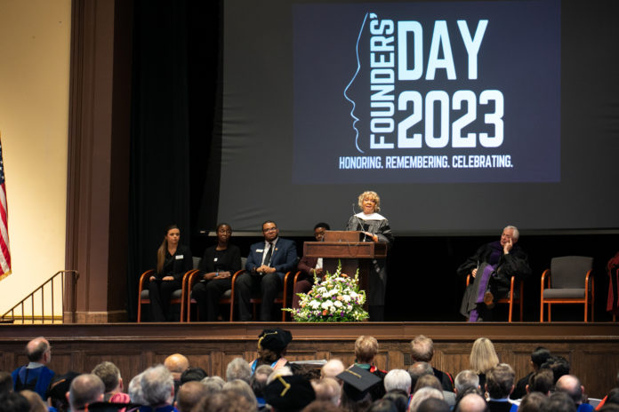 A pearlie toliver, wearing academic regalia, stands at a podium and speaks. behind here, the words Founders' Day 2023 are projected on a screen.