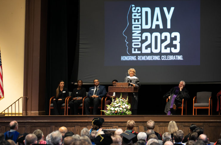 A pearlie toliver, wearing academic regalia, stands at a podium and speaks. behind here, the words Founders' Day 2023 are projected on a screen.