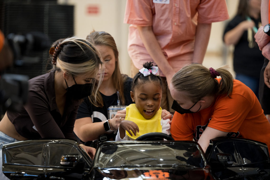 a child sits in a toy car while three young people help fit her in it.