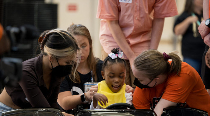 Mercer to host ninth ‘Go Baby Go’ toy car build for kids with limited mobility a child sits in a toy car while three young people help fit her in it.