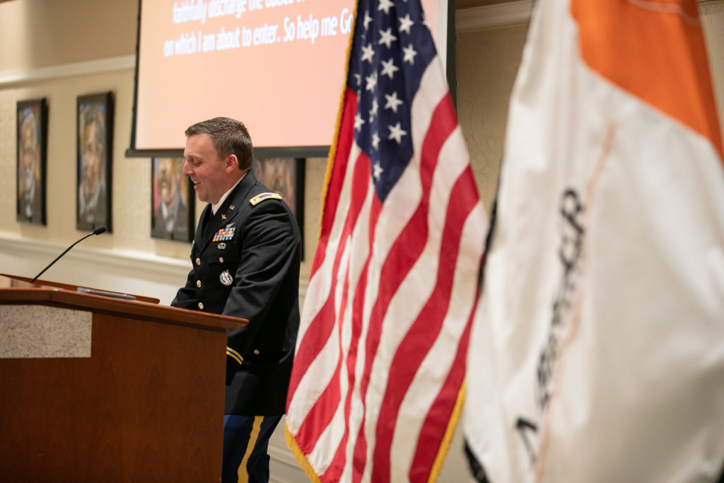 lt. col. pitchford, wearing a military officer uniform, speaks at a podium. an american flag and a mercer flag are in the foreground.