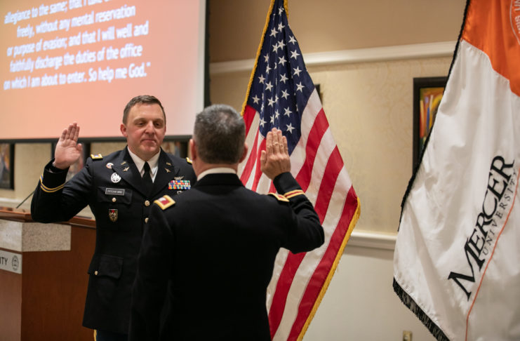 two men in military officer uniforms raise their right hands while standing in front of an american flag and a mercer flag