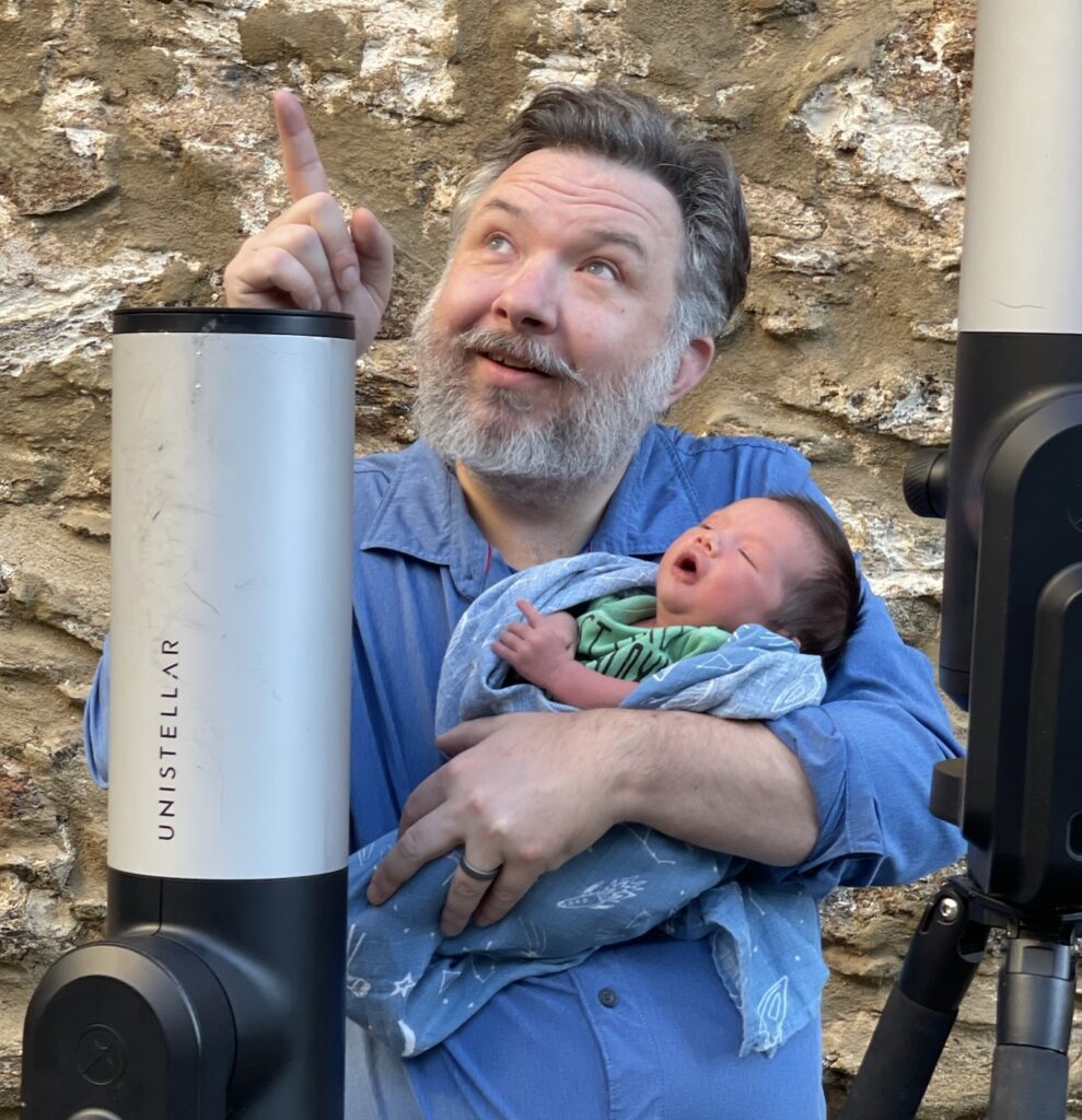 Justus Randolph points to the sky with one hand while holding his newborn son in the other, with a telescope in front of them.